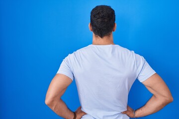 Young hispanic man standing over blue background standing backwards looking away with arms on body
