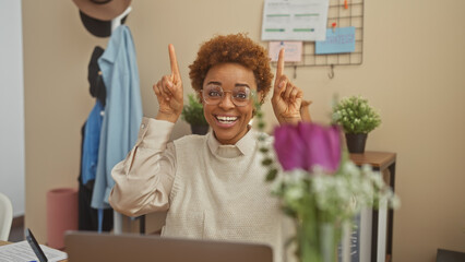 Cheerful african american woman pointing upwards, smiling in a cozy home office setup.