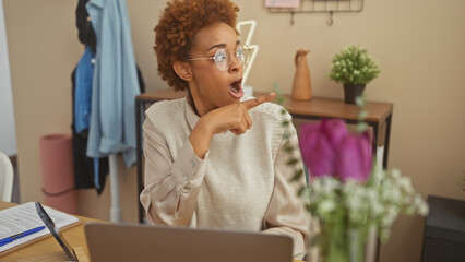African american woman pointing excitedly in a home office setting with a laptop and flowers in the...