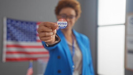 African american woman in a blue blazer showing 'i voted' sticker with us flag background indoors