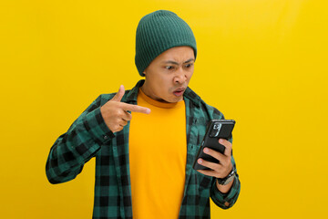 An enraged Asian man, dressed in a beanie hat and casual shirt, points at his mobile phone while visibly reacting to bad news, showing intense anger and fury, while standing against yellow background