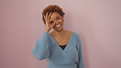 A smiling black woman making an okay sign over her eye, against a pink background.