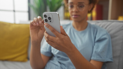 African american woman uses smartphone, sitting indoors on a couch in a well-lit living room, exhibiting casual, modern lifestyle.