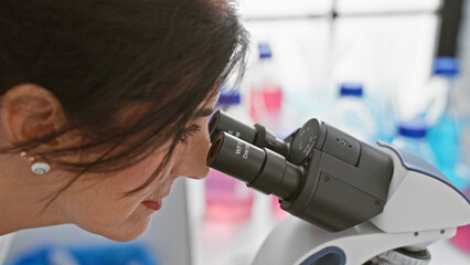 A brunette woman examines samples through a microscope in a laboratory setting with blurred...