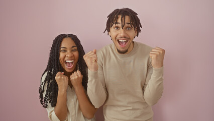 A cheerful young couple celebrates with fists raised against a plain pink background.