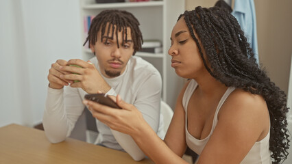 A displeased woman and a man sit indoors at home, suggesting relationship issues as she looks at her smartphone.