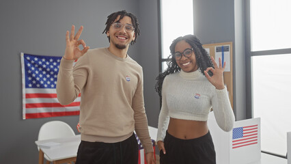 A smiling couple with 'i voted' stickers makes the ok sign indoors, with an american flag in the...