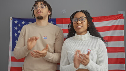 African american couple with 'i voted' stickers applauding in a room with a us flag background.