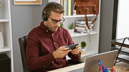 Professional hispanic man with headphones using smartphone in modern office.