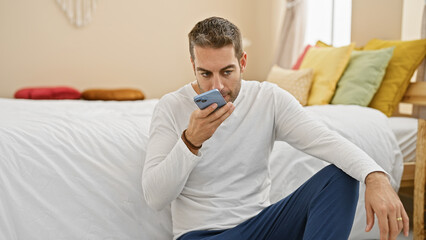 Fototapeta premium A handsome young adult hispanic man with a beard sending a voice message on his smartphone at home in his bedroom