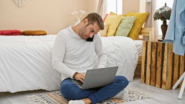 A young bearded hispanic man multitasking with a laptop and phone in the cozy bedroom of a modern apartment.