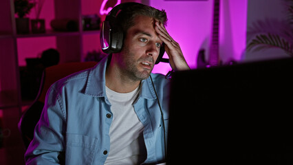 Hispanic man with headphones in a dark room looks stressed while using a computer at night.