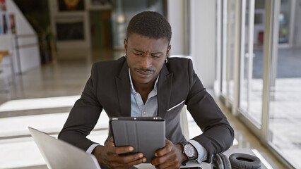 A focused african american man in formal attire uses a tablet at his modern office space.