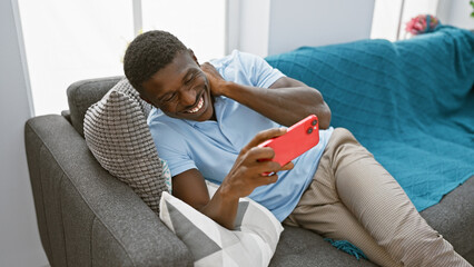 Handsome african american man laughing while using smartphone on couch at home