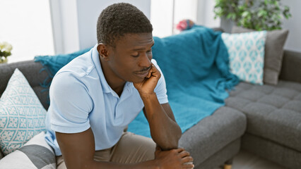 Pensive african man sitting indoors in a modern apartment living room contemplating.
