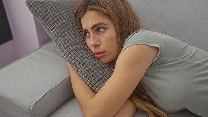 A beautiful, contemplative young caucasian woman lounges indoors, her gaze distant, resting thoughtfully on a sofa pillow.