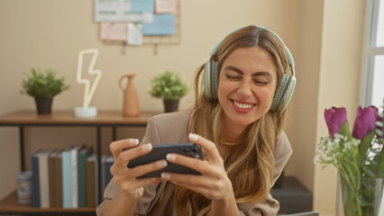 Smiling young caucasian woman wearing headphones plays a mobile game at home.