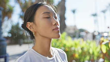 A serene young asian woman with eyes closed, enjoying the calm ambiance of a sunlit urban park.