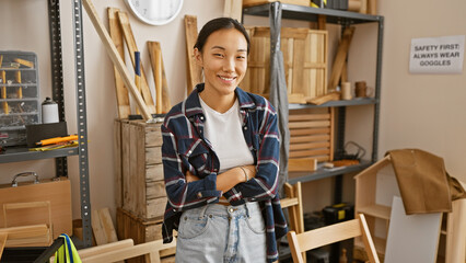 Confident asian woman with crossed arms standing in a carpentry workshop surrounded by wood and...