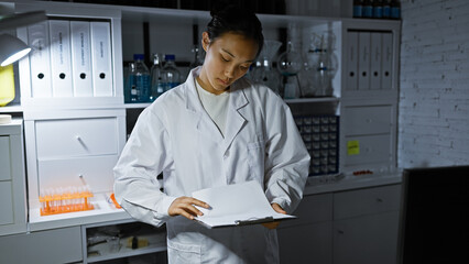 A focused asian woman scientist in a lab coat analyzing documents in a modern laboratory setting.