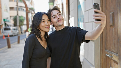 An interracial couple, a woman and a man, take a selfie together on a sunny urban street.