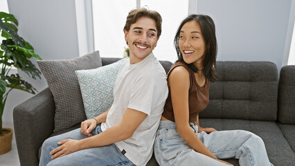 An interracial couple sits together smiling in a cozy living room, embodying a modern, loving...
