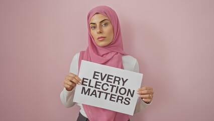 A confident young woman in a hijab holding a sign saying 'every election matters' against a pink...