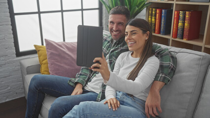 Smiling hispanic couple taking a selfie with a tablet while sitting on a sofa in their modern...