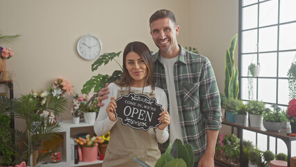 Fototapeta premium Smiling man and woman holding an 'open' sign in a vibrant indoor flower shop filled with various plants.
