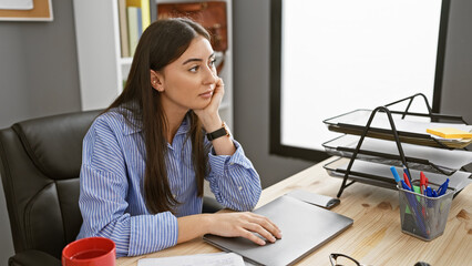 Pensive young hispanic woman sitting at her office desk, looking away thoughtfully, embodying a...