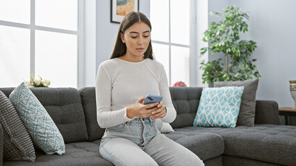 Hispanic woman in white sweater casually using a smartphone while seated on a gray couch indoors