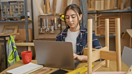 A focused young hispanic woman works on a laptop in a cluttered woodworking workshop, surrounded by...