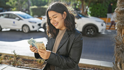 A smiling young woman examines brazilian reais on a sunny urban street, reflecting everyday...
