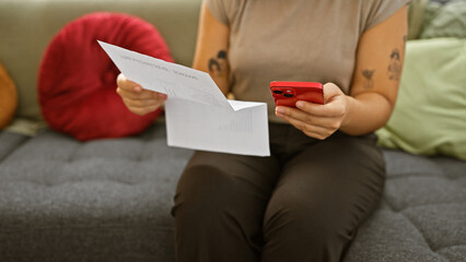 Hispanic woman using smartphone and reading document indoors, portraying a modern adult in a living...