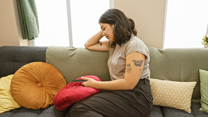 Young hispanic woman using smartphone at home, with tattoos visible, sitting on a sofa surrounded...