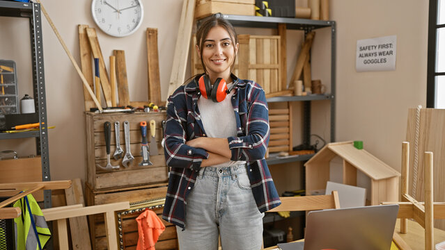 Confident hispanic woman with crossed arms stands in a carpentry workshop, sporting safety headphones.