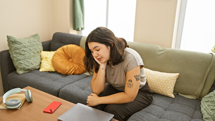 A thoughtful young hispanic woman with tattoos sitting on a grey sofa in a cozy living room with colorful pillows, headphones, and laptop.