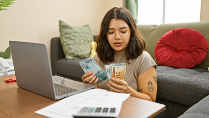 Young hispanic woman counts brazilian reais in a cozy apartment's living room with a laptop on the...