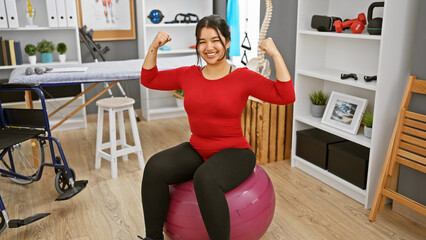 Hispanic woman exercises with a fitness ball in a rehab clinic's therapy room.