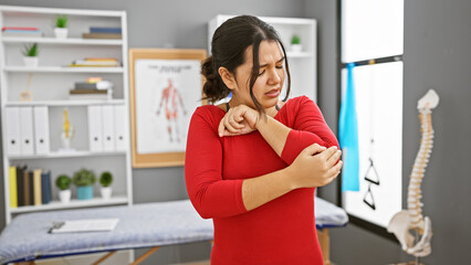 Hispanic woman in pain holding elbow at physical therapy clinic, showing discomfort and injury.