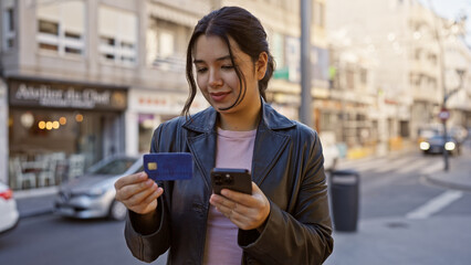 A young hispanic woman examines her credit card while holding a smartphone on a sunny city street.