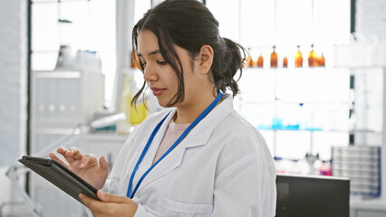 A young hispanic woman in a white lab coat uses a tablet in a bright medical laboratory, conveying...