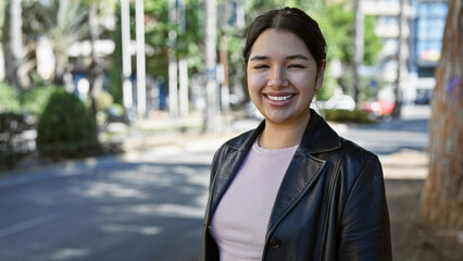 Smiling young hispanic woman in leather jacket standing outdoors in a sunny city park.