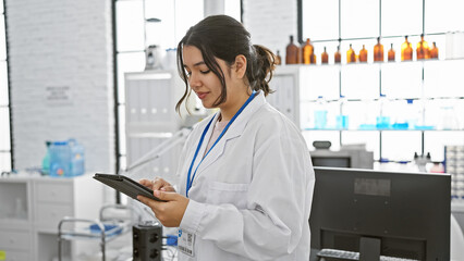 Young hispanic woman in white lab coat using tablet in modern laboratory setting, focused and...