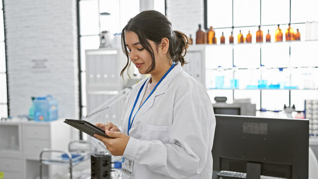 Young hispanic woman in white lab coat using tablet in modern laboratory setting, focused and professional.