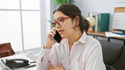 Hispanic woman talking on phone in office setting, portraying a professional adult in an indoor work environment.