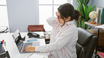 A professional hispanic woman experiences neck pain while working on a laptop in a modern office...