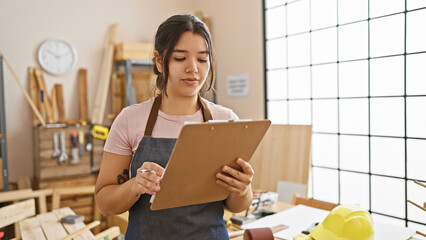 Hispanic woman reviewing plans in a workshop with woodworking tools, safety gear, and warm lighting.