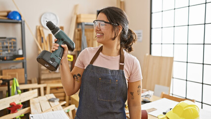 Hispanic woman smiles holding drill in carpentry workshop, with safety glasses and apron