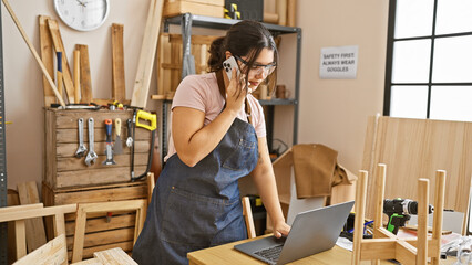 Young hispanic woman in apron using laptop and phone in a carpentry workshop